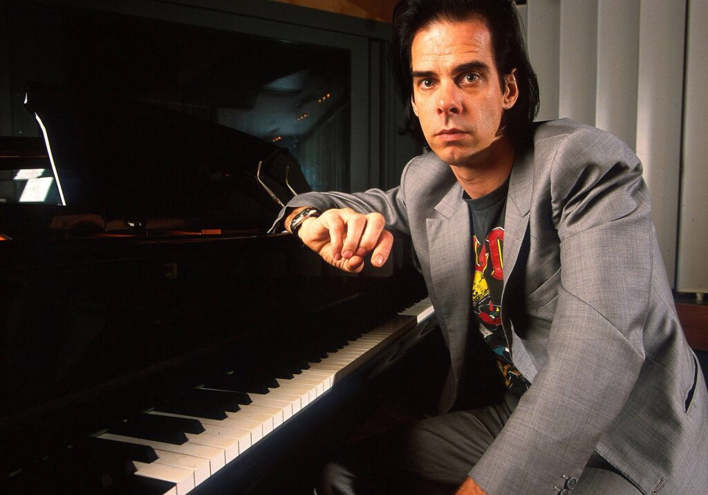 Nick Cave sitzt an einem Flügel, hat den rechten Arm oben auf dem Instrument aufgestützt und wendet sich zur Kamera.